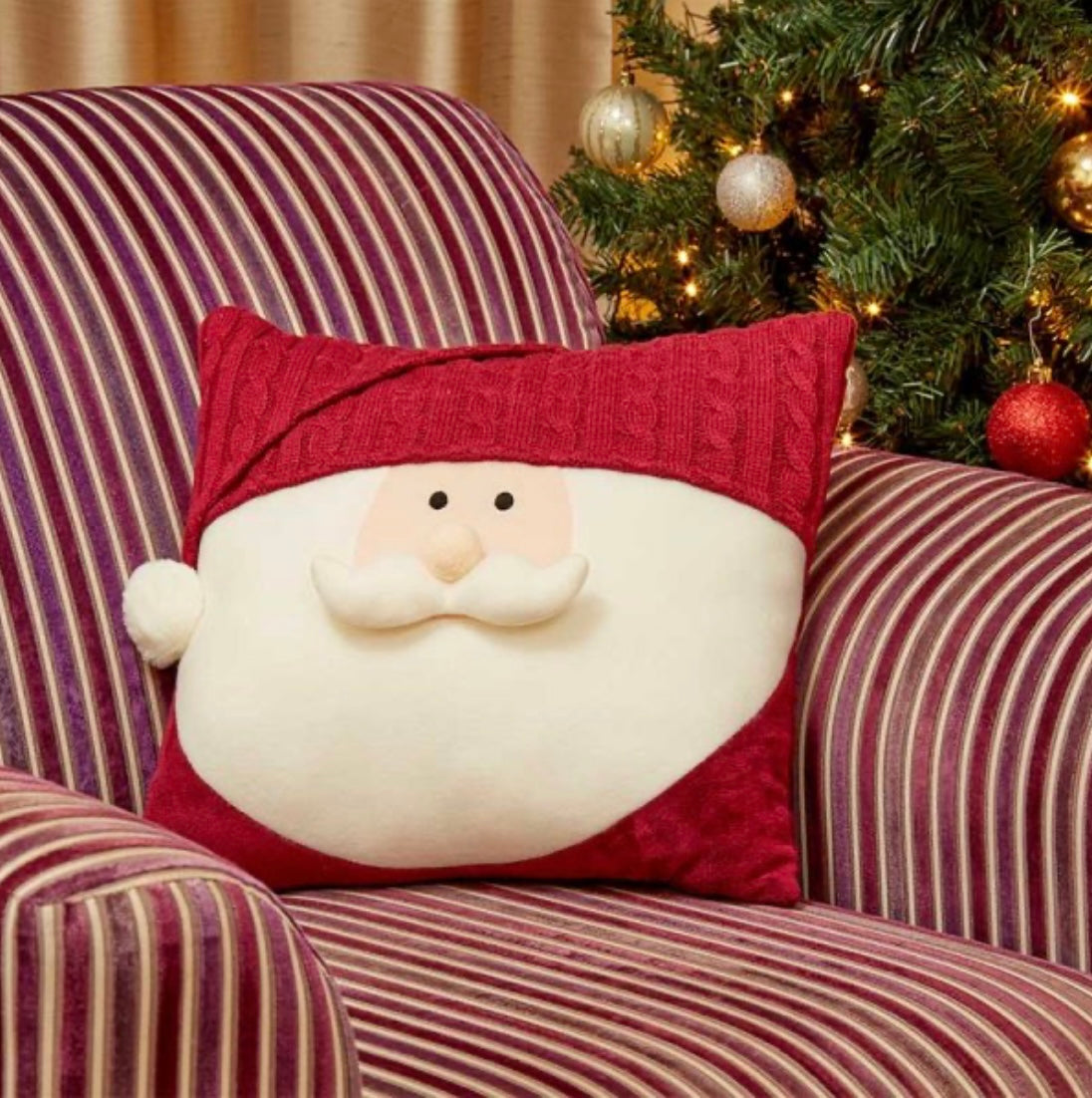 A festive Santa-shaped cushion with a red trim and a white pom-pom, placed on a striped chair with a Christmas tree and lights in the background.