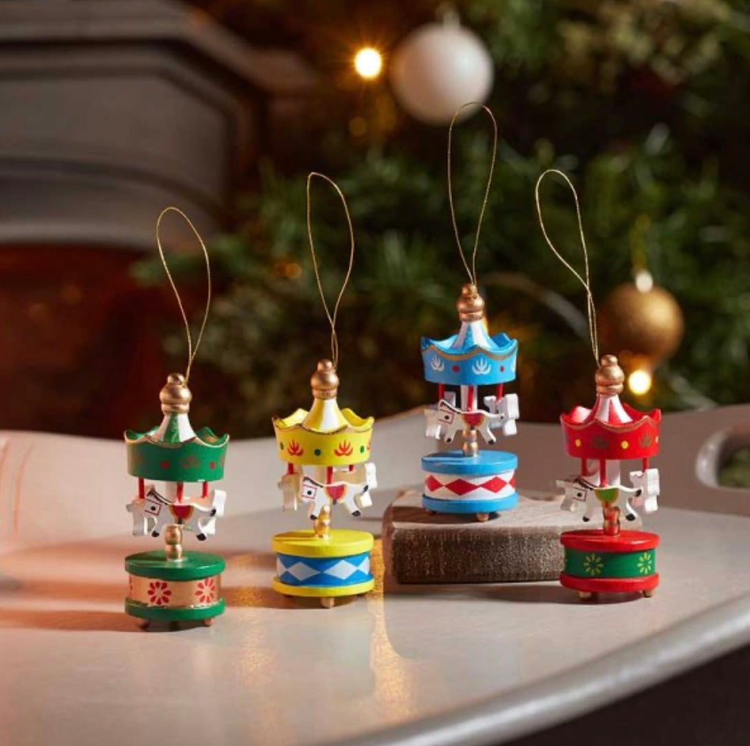 A set of four hand-painted Christmas carousel decorations, each with a different color scheme, displayed on a flat surface with a blurred Christmas tree and lights in the background.