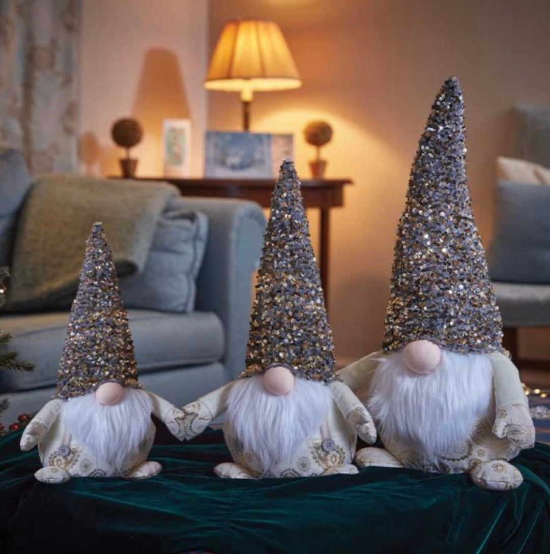 Three jumbo-sized decorative gnomes with glittery hats, seated indoors on a green cloth.