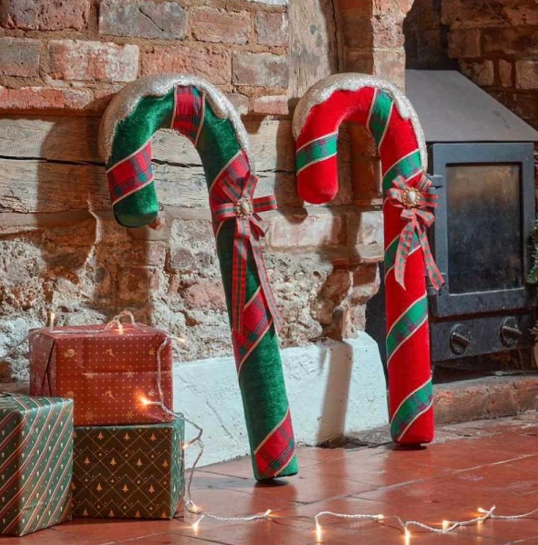 Two candy cane harlequin ornaments in burgundy and emerald colors with green and red stripes, standing upright next to a brick wall and a decorated gift box.