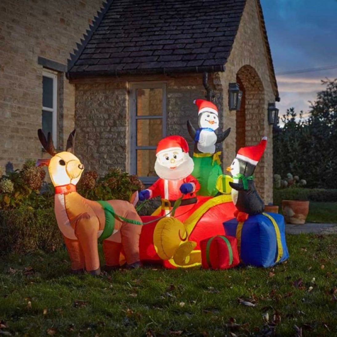 A self-inflating Santa's sleigh ride decoration featuring a red sleigh with Santa and reindeer on a grassy area in front of a house at dusk.