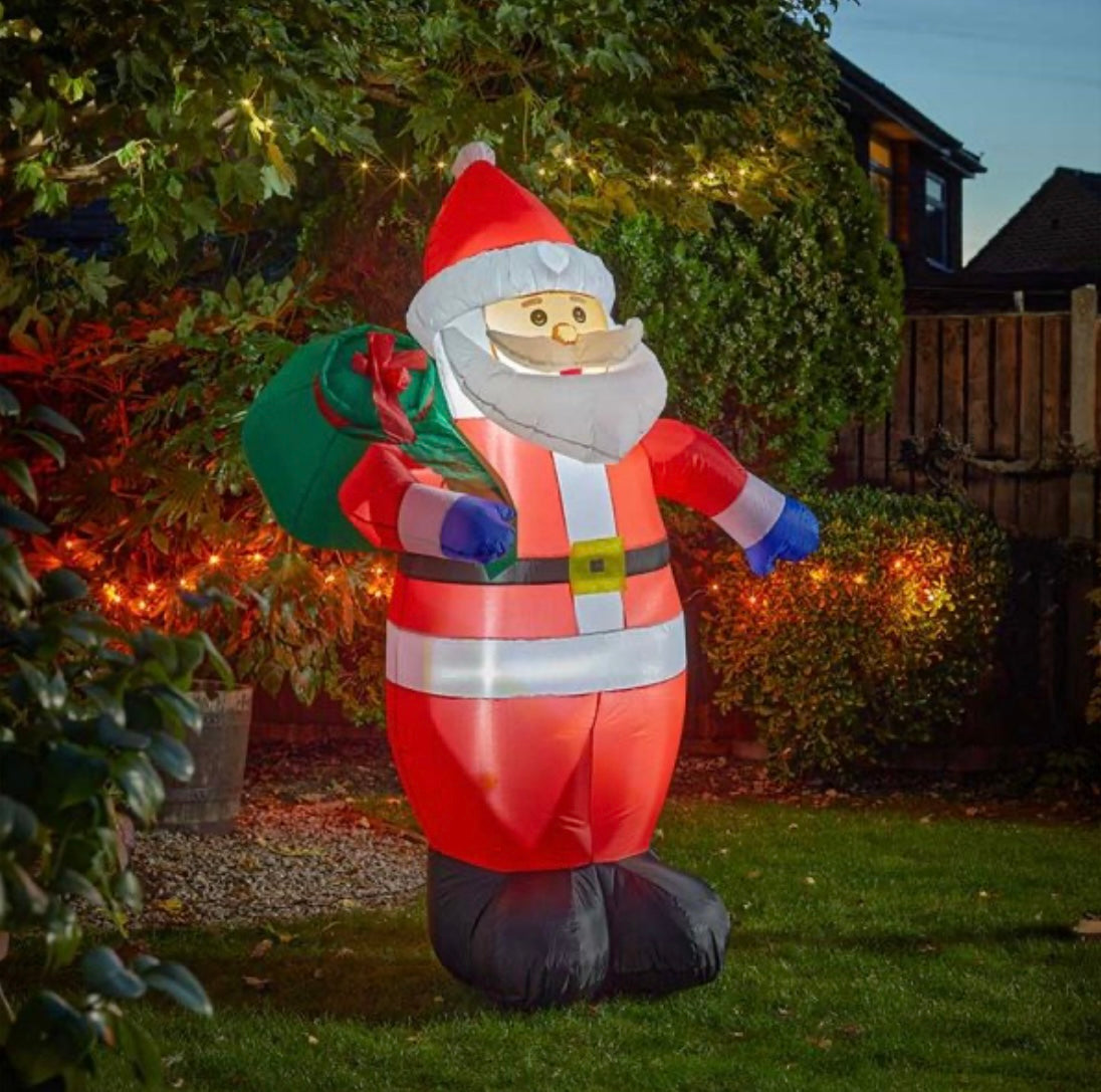 An inflatable Santa Claus figure with a red suit and a green Christmas tree, standing in a garden at night with house lights and decorations in the background.