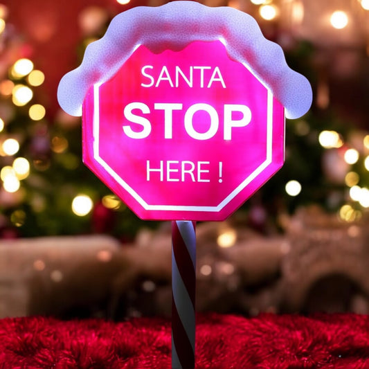 A red 'Santa Stop Here' sign with a white border and a snowman illustration, placed indoors with Christmas decorations around.