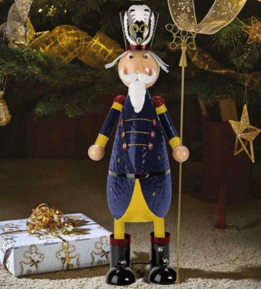 A decorative nutcracker standing next to a Christmas tree, wearing a blue jacket, yellow pants, and black boots, with a golden hat and star-shaped embellishments.