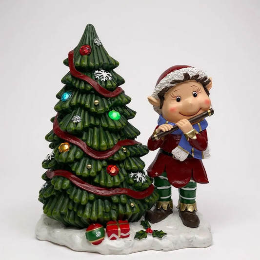 Elf figurine playing a flute next to a decorated Christmas tree on a white background
